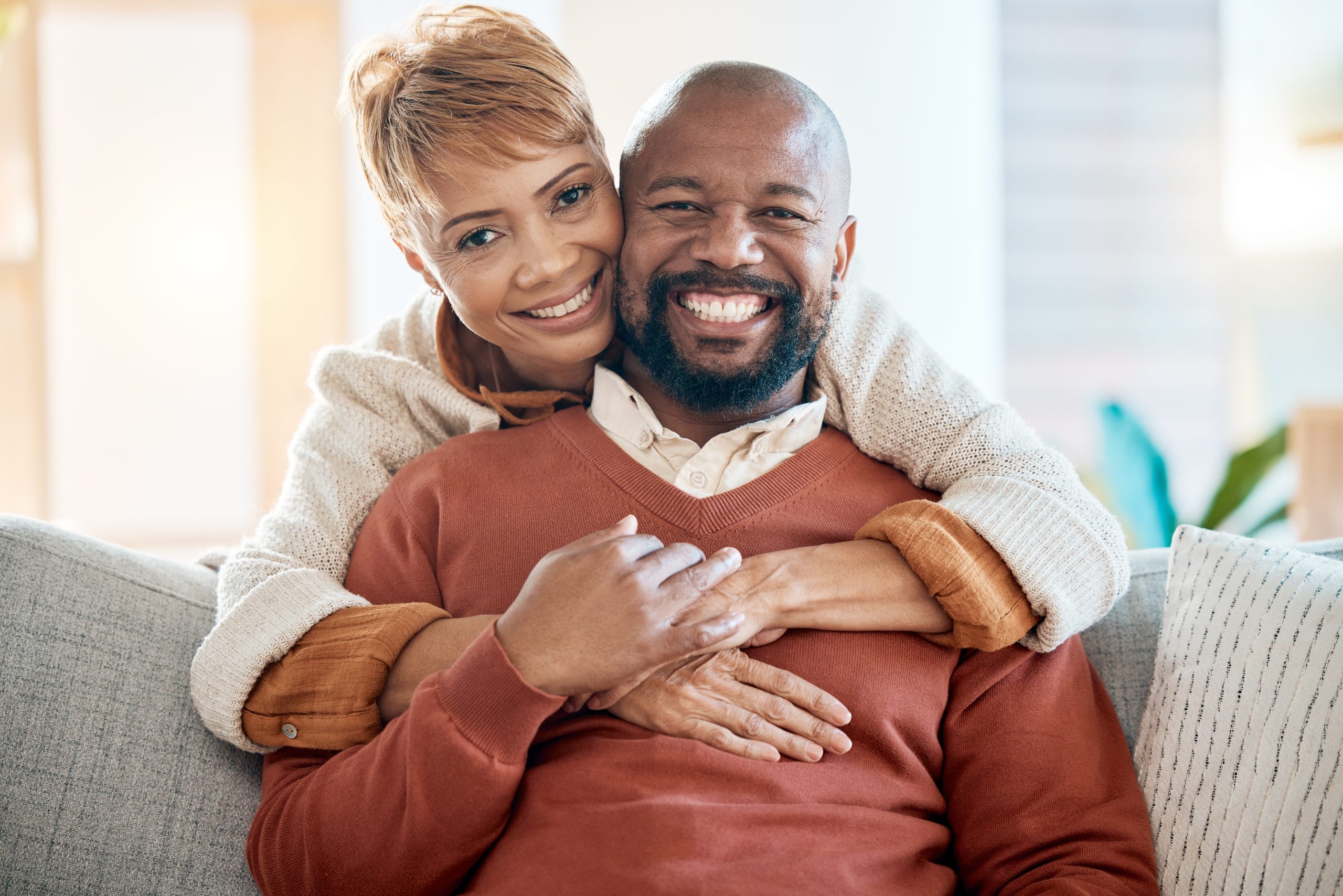 A woman hugs a smiling man from behind as they sit together on a couch, both looking at the camera.