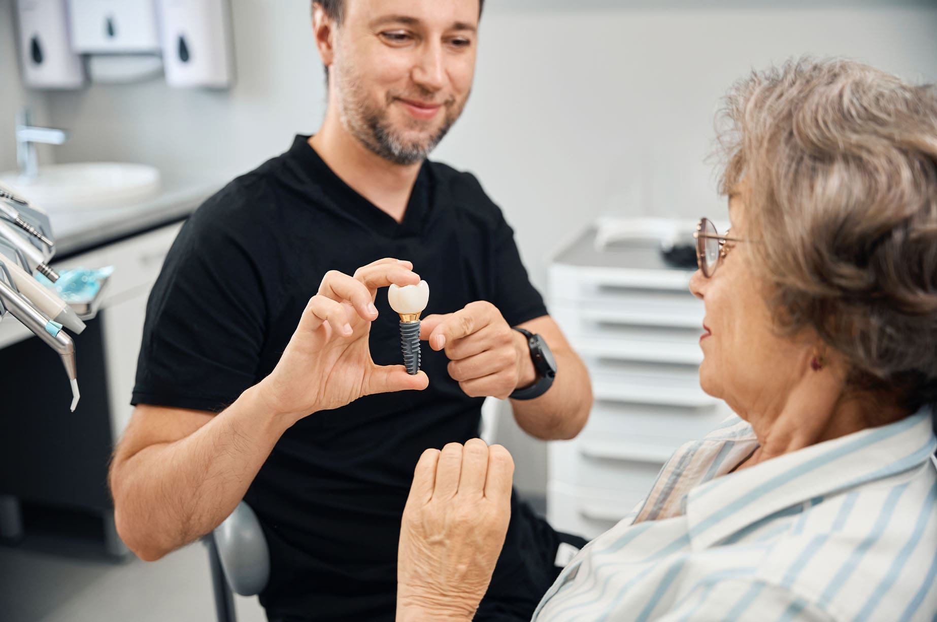 A dentist shows a dental implant model to an older woman while sitting in a dental clinic office.