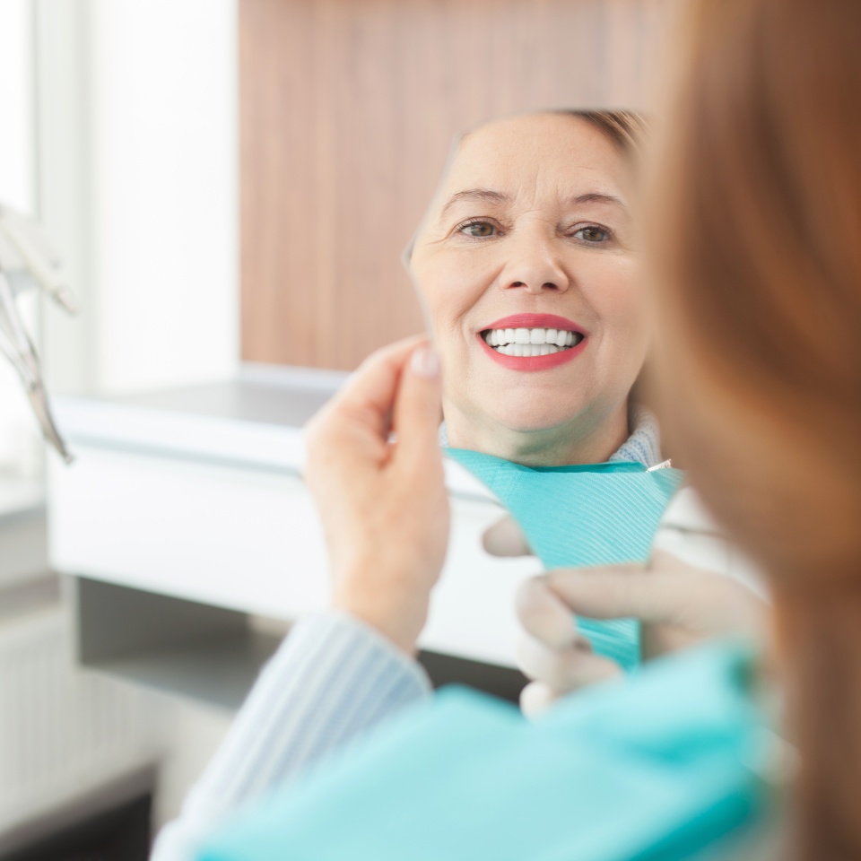 Pretty senior woman is visiting her dentist. She is holding a mirror and looking at her teeth with joy. The woman is smiling happily