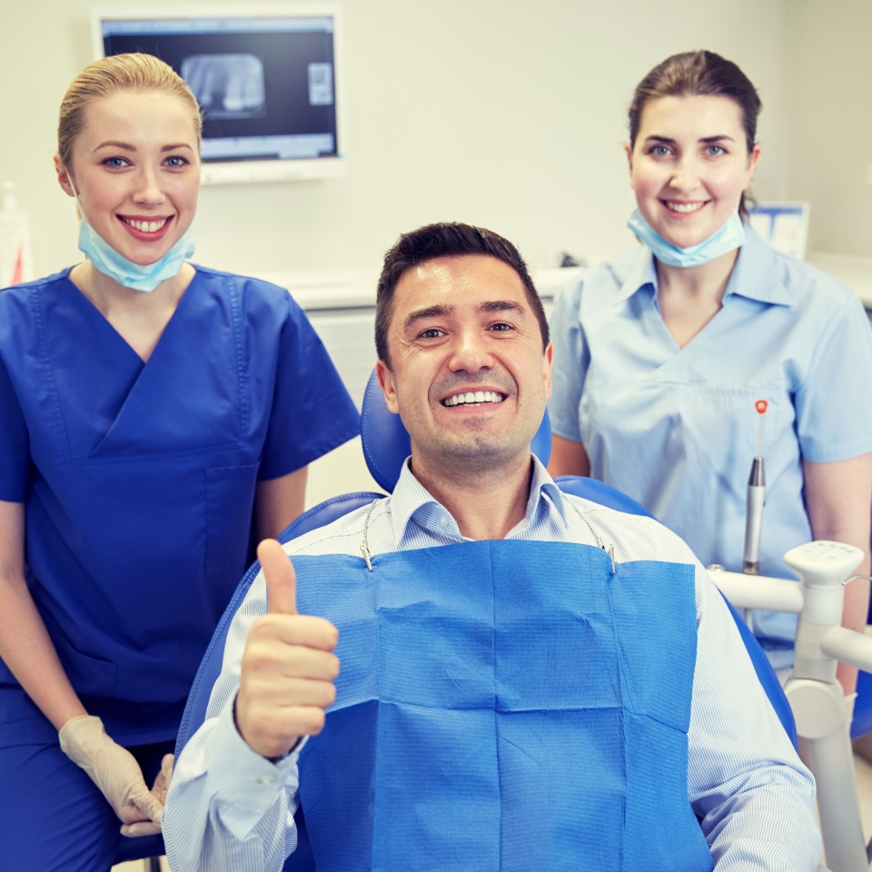 people, medicine, stomatology, gesture and health care concept - happy female dentist with assistant and man patient showing thumbs up at dental clinic office