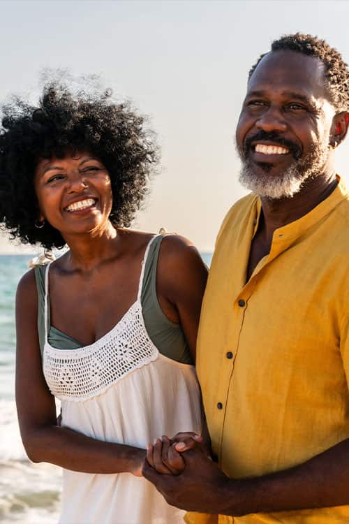 A smiling man and woman stand close together on a beach, holding hands, with the ocean and sky in the background.