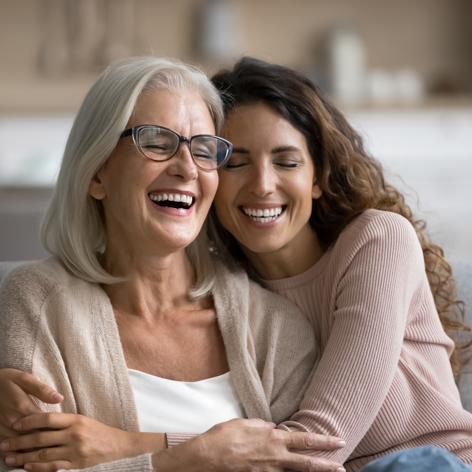 Happy excited senior mom and adult daughter woman having fun at home, hugging with heads touch, sitting on sofa, laughing with closed eyes, enjoying funny talk, leisure, family affection