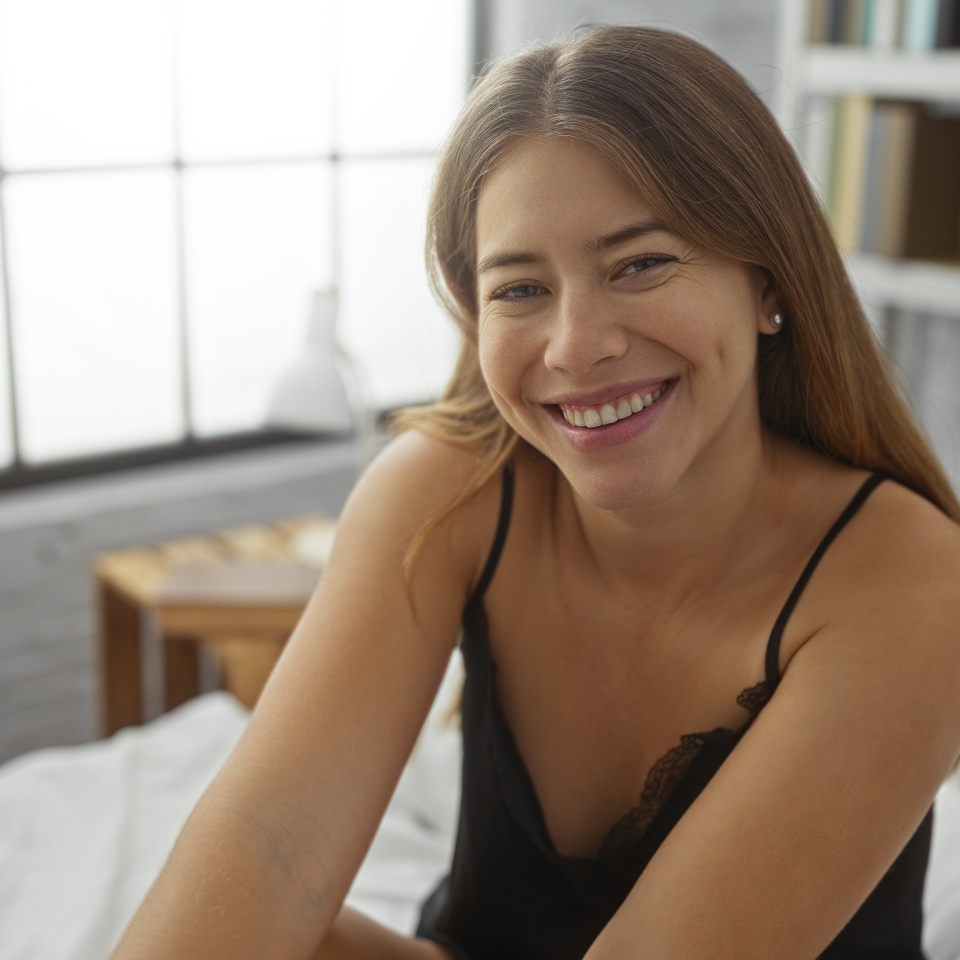 Smiling woman sitting on bed in cozy bedroom with bookshelves and natural light, showcasing an attractive and relaxed atmosphere at home.